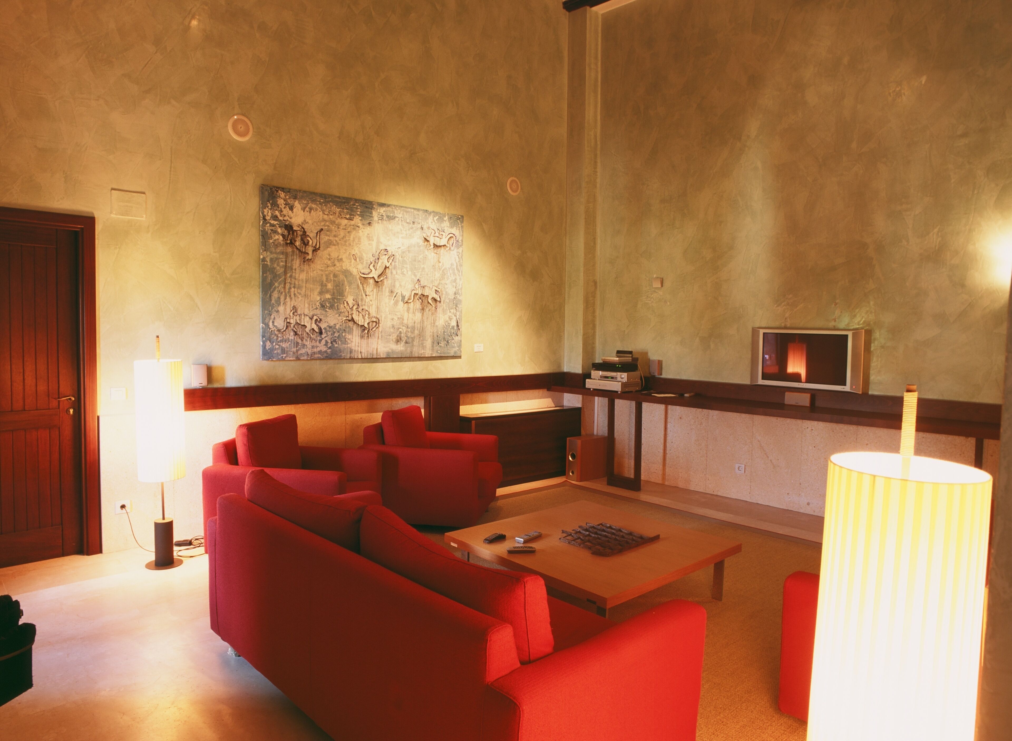 Una sala de estar con sofás rojos, una mesa de centro de madera, un cuadro abstracto grande, un televisor y una lámpara.