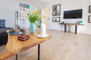 A living room with a wooden coffee table, a vase with flowers, a white candle, and a flat-screen TV mounted on the wall.