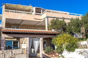 A two-story building with a balcony, stone facade, and a small garden with yellow flowers.
