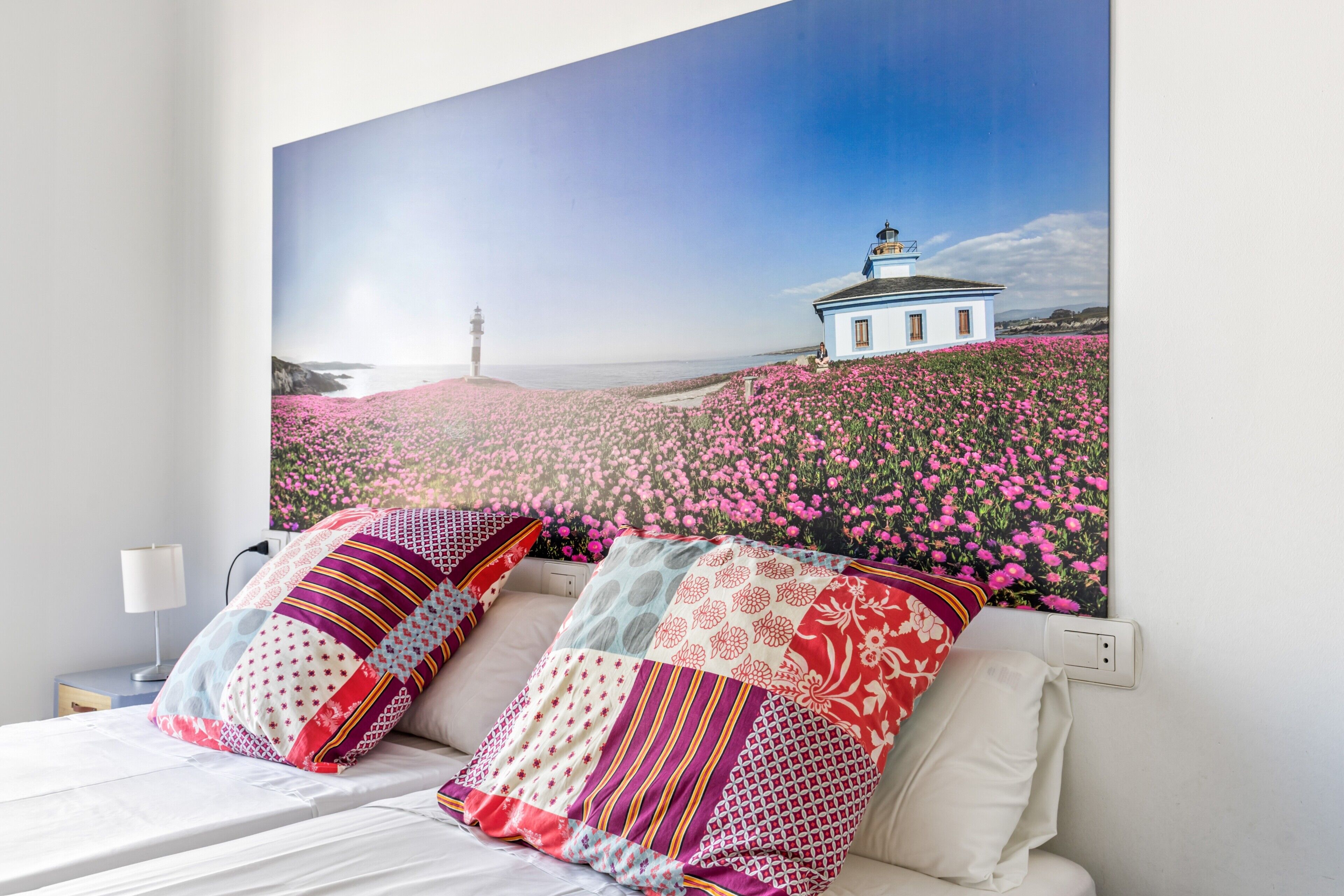 A bedroom with a large bed, two pillows, a nightstand, a lamp, and a wall-mounted painting of a coastal scene.