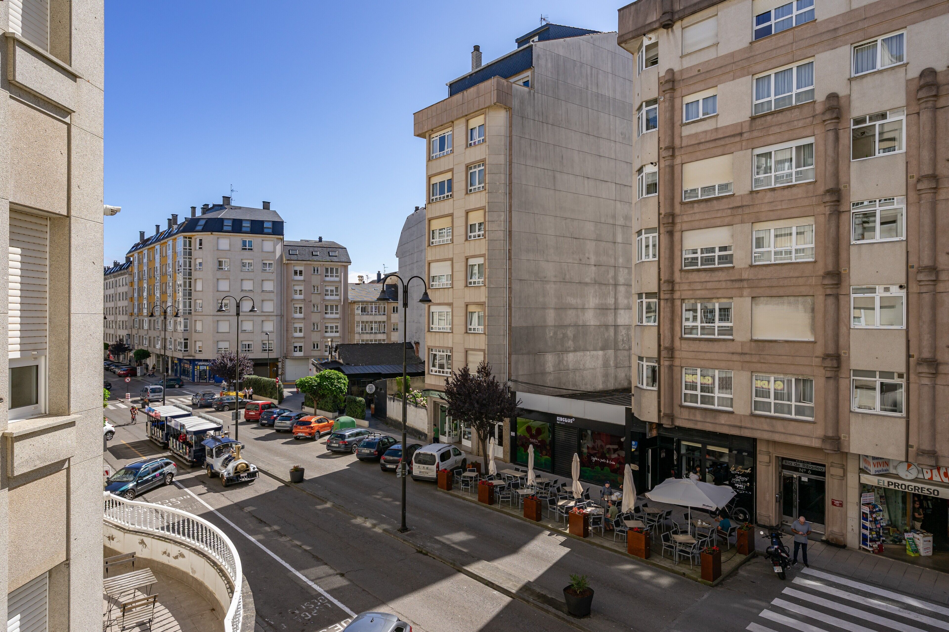 Eine Stadtstraße mit Gebäuden, parkenden Autos und Außenbereichsgastronomie.