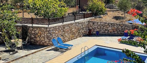 A rectangular swimming pool with a stone wall and a wooden fence, surrounded by lounge chairs and umbrellas.