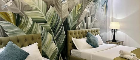 A hotel room with two beds, a mural of tropical leaves, a chandelier, and a bedside table with a lamp.