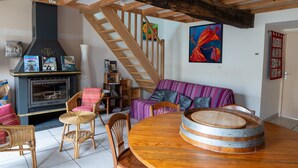 Un salon chaleureux avec une cheminée, des poutres apparentes, un escalier et un tableau aux couleurs vives.