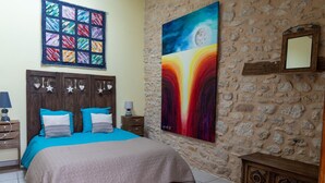 Une chambre à coucher avec un lit, une table de chevet, une commode, une courtepointe et un tableau accroché à un mur en pierre.