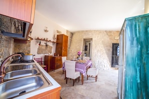 A kitchen with a gas stove, sink, and wooden cabinets. There is a dining table with chairs and a tablecloth. The walls are made of stone, and there is a window with a view of the outside.