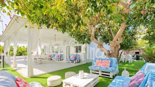 An outdoor seating area with white furniture under a white canopy, surrounded by lush greenery and a large tree.