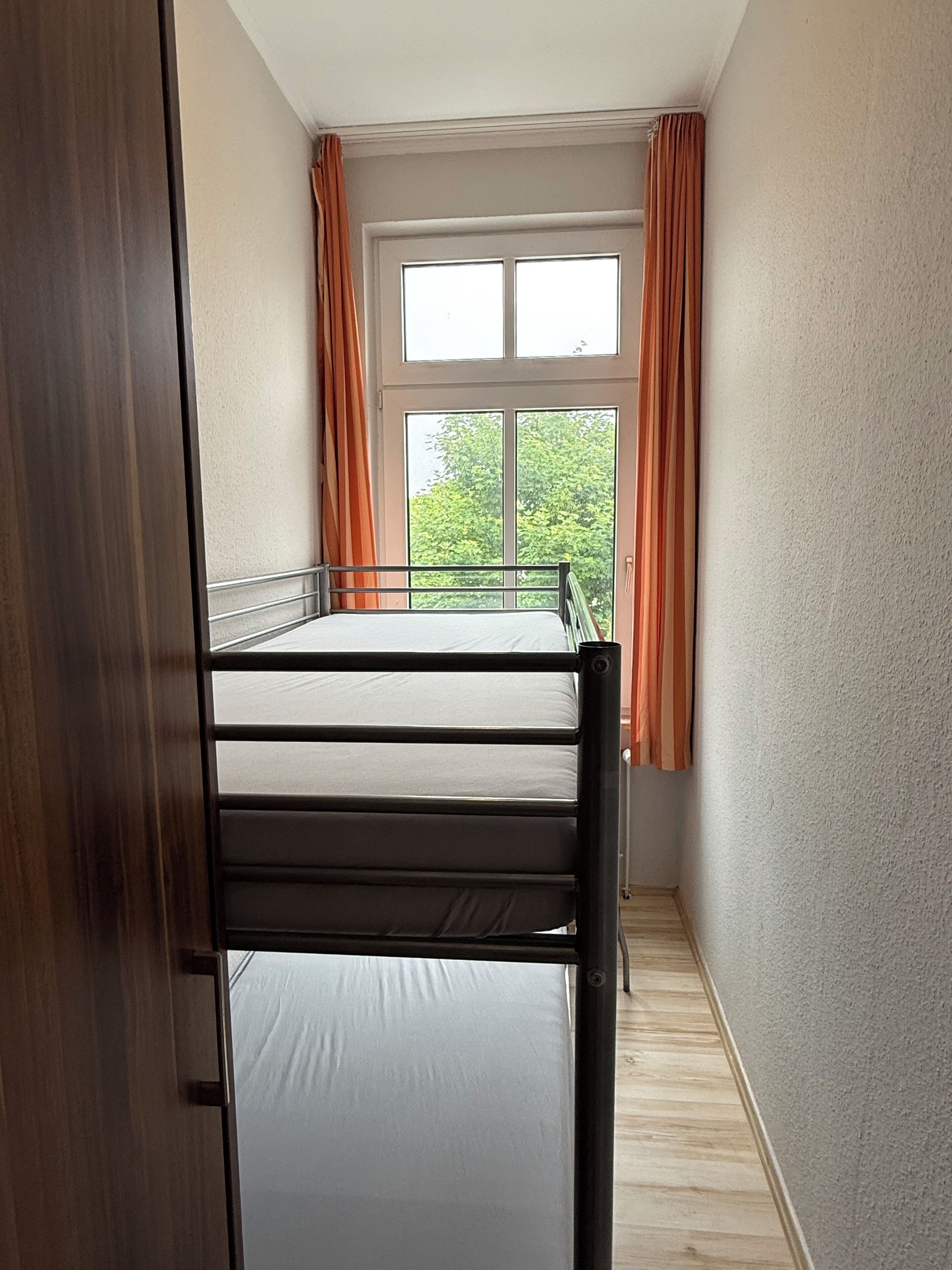 Quarto pequeno com uma cama de solteiro, janela e cortinas, piso de madeira e porta.