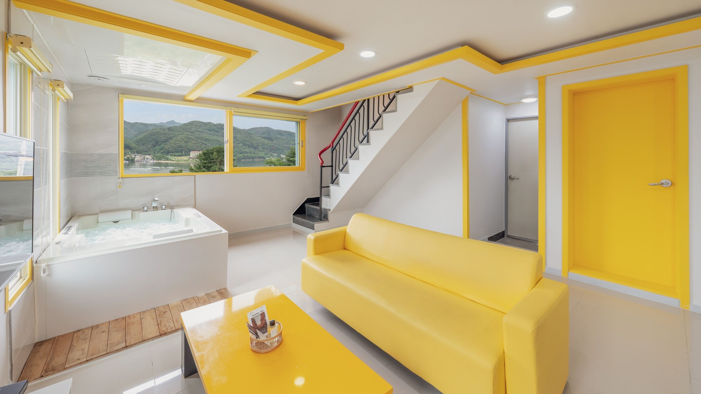 A modern living room with a yellow sofa, a large window with a view of greenery, a staircase, and a bathroom with a bathtub.