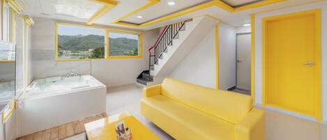 A modern living room with a yellow sofa, a large window with a view of greenery, a staircase, and a bathroom with a bathtub.
