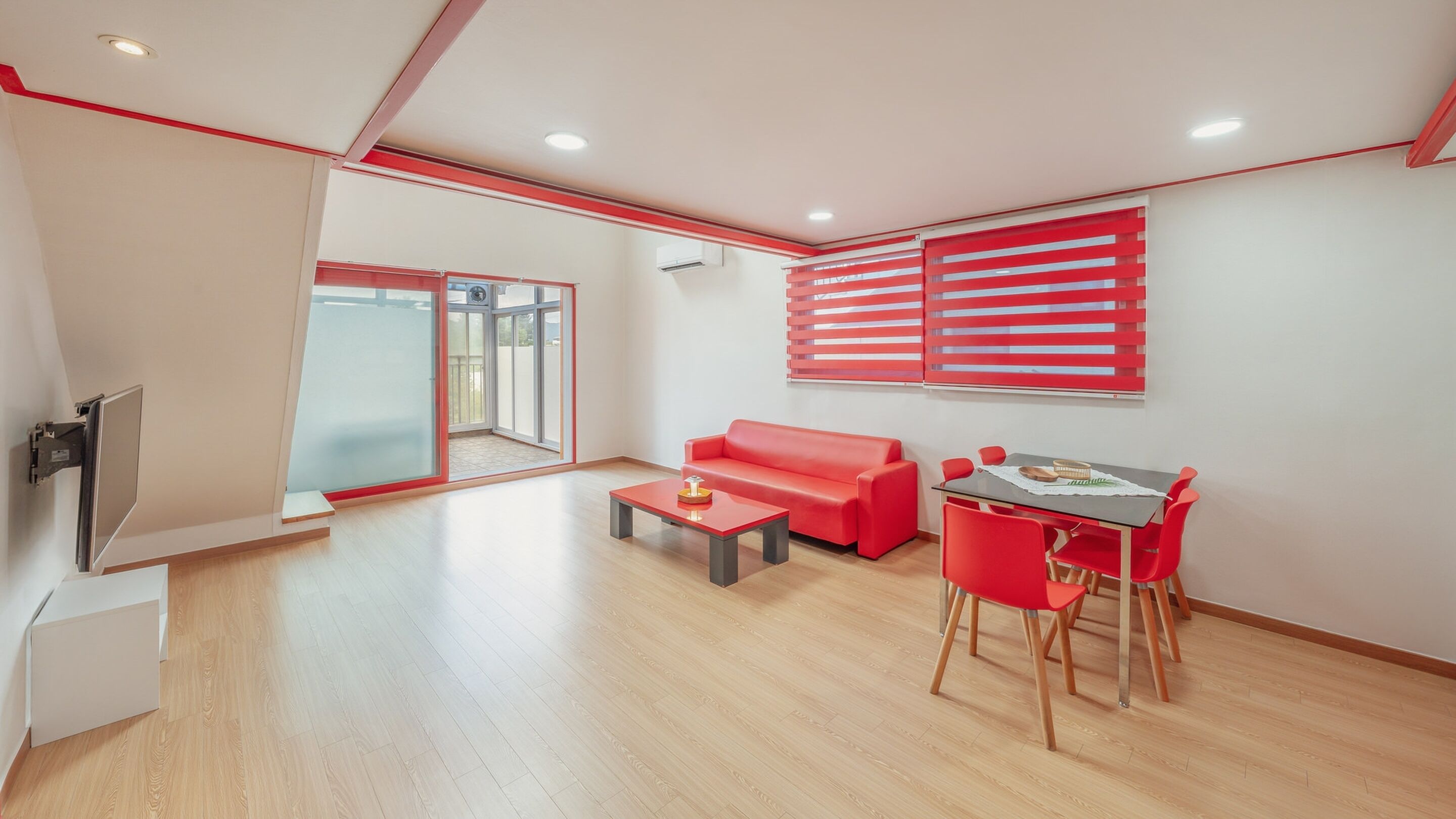 A modern living room with a red sofa, a small table, and a red chair. There is a sliding glass door leading to a balcony.