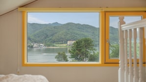 Quarto com uma grande janela com vista para um lago, um edifício na outra margem e uma colina arborizada.