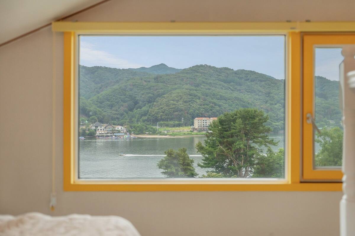 Ein Zimmer mit großem Fenster mit Blick auf einen See, ein Gebäude gegenüber dem Wasser und ein bewaldeter Hügel.