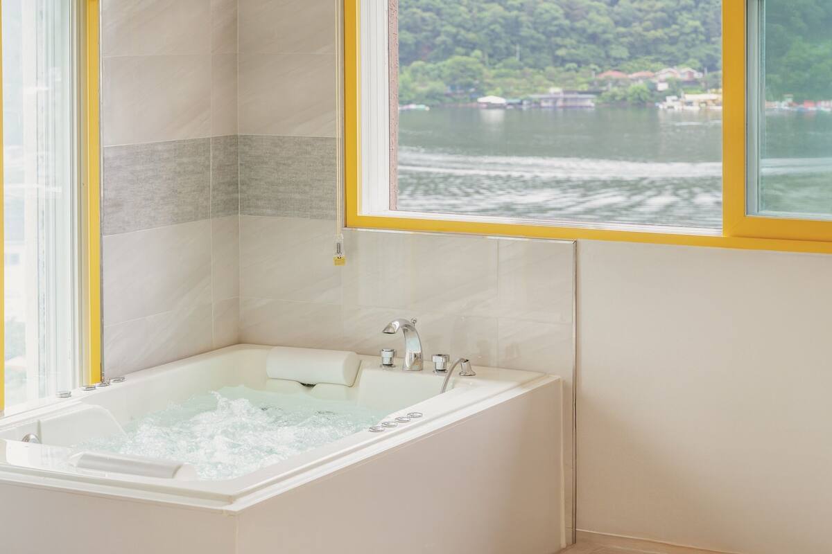 Ein modernes Badezimmer mit einer Badewanne, einem Fenster mit gelbem Rahmen und Blick auf einen See mit Booten.