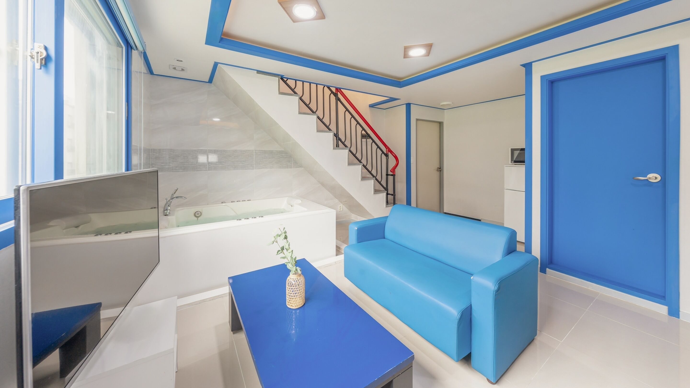 A modern hotel room with a blue sofa, a small table with a plant, a bathtub, and a staircase with red railings.