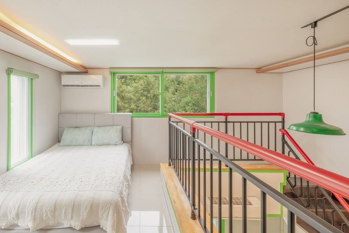 Ein Schlafzimmer mit einem Bett, einem Fenster, einer Treppe und einer grünen Pendelleuchte.