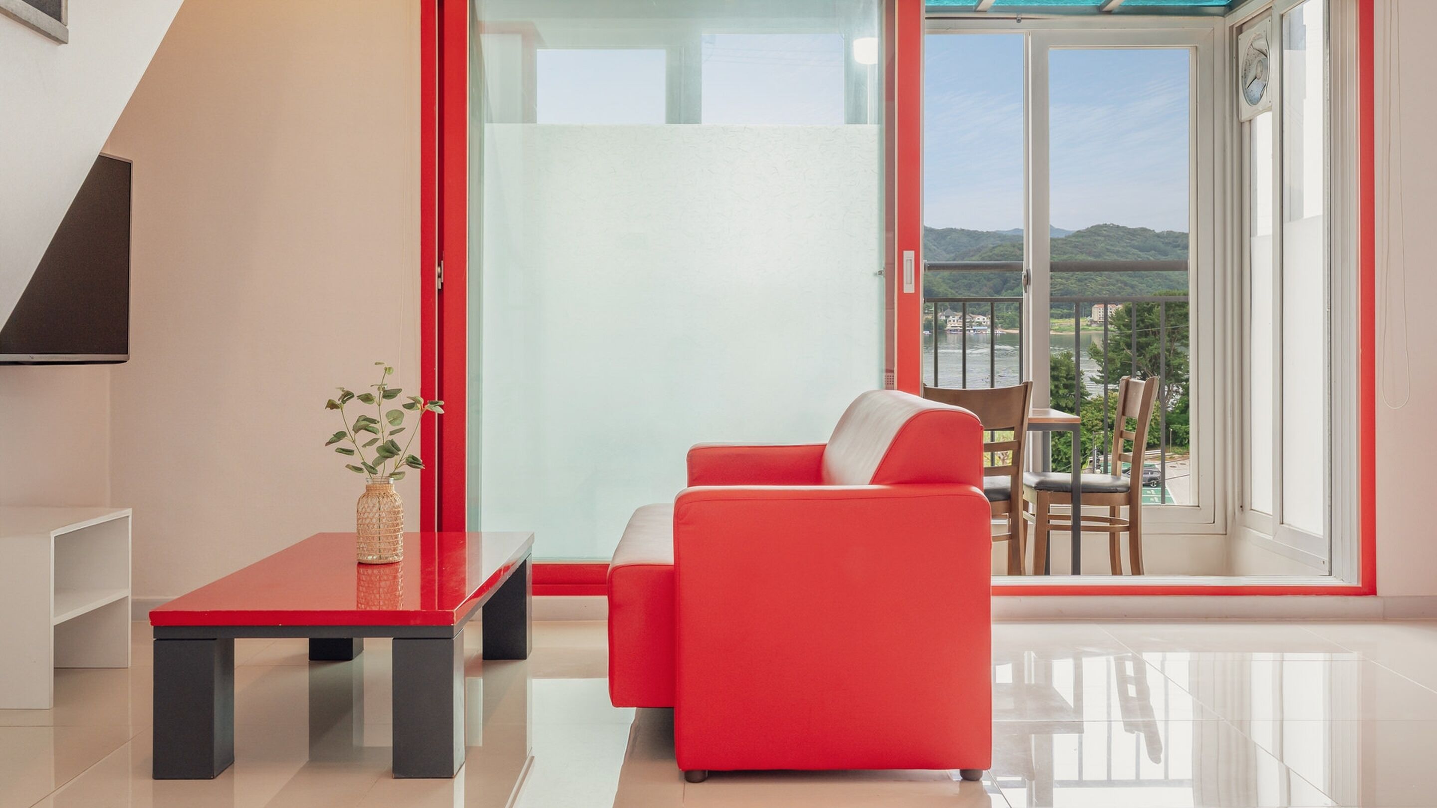 Een moderne woonkamer met een rode bank, een glazen tafel en uitzicht op een balkon met buitenmeubilair.
