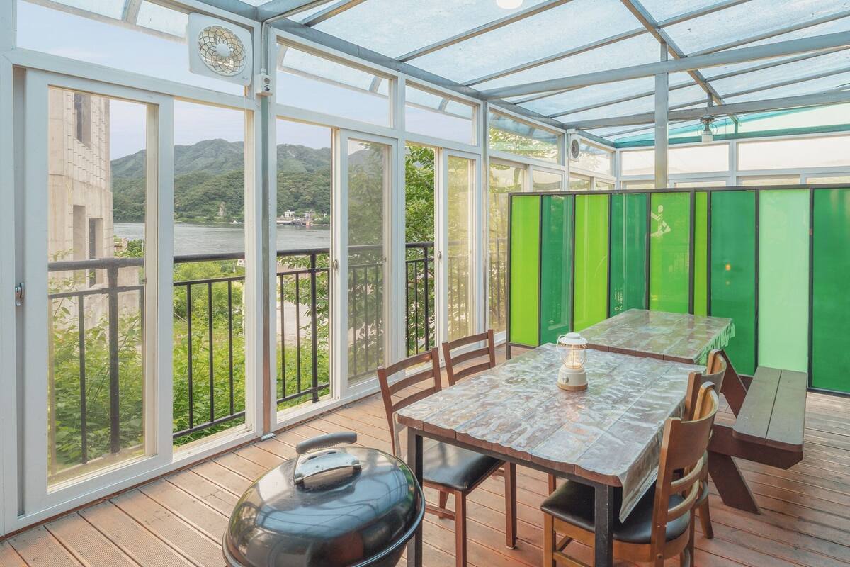 Eine überdachte Terrasse mit Tisch und Stühlen, einem Grill und Blick auf einen Fluss und Hügel.