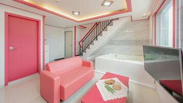 Una sala de estar moderna con un sofá rosa, una mesa de centro de vidrio, un televisor de pantalla plana y una escalera que lleva a un baño con bañera.
