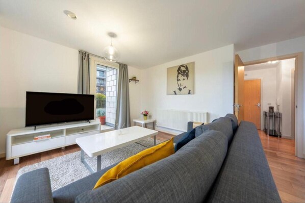 Un salon avec un canapé gris, une table basse blanche, un téléviseur à écran plat posé sur un meuble blanc, une plante en pot et un tableau accroché au mur.