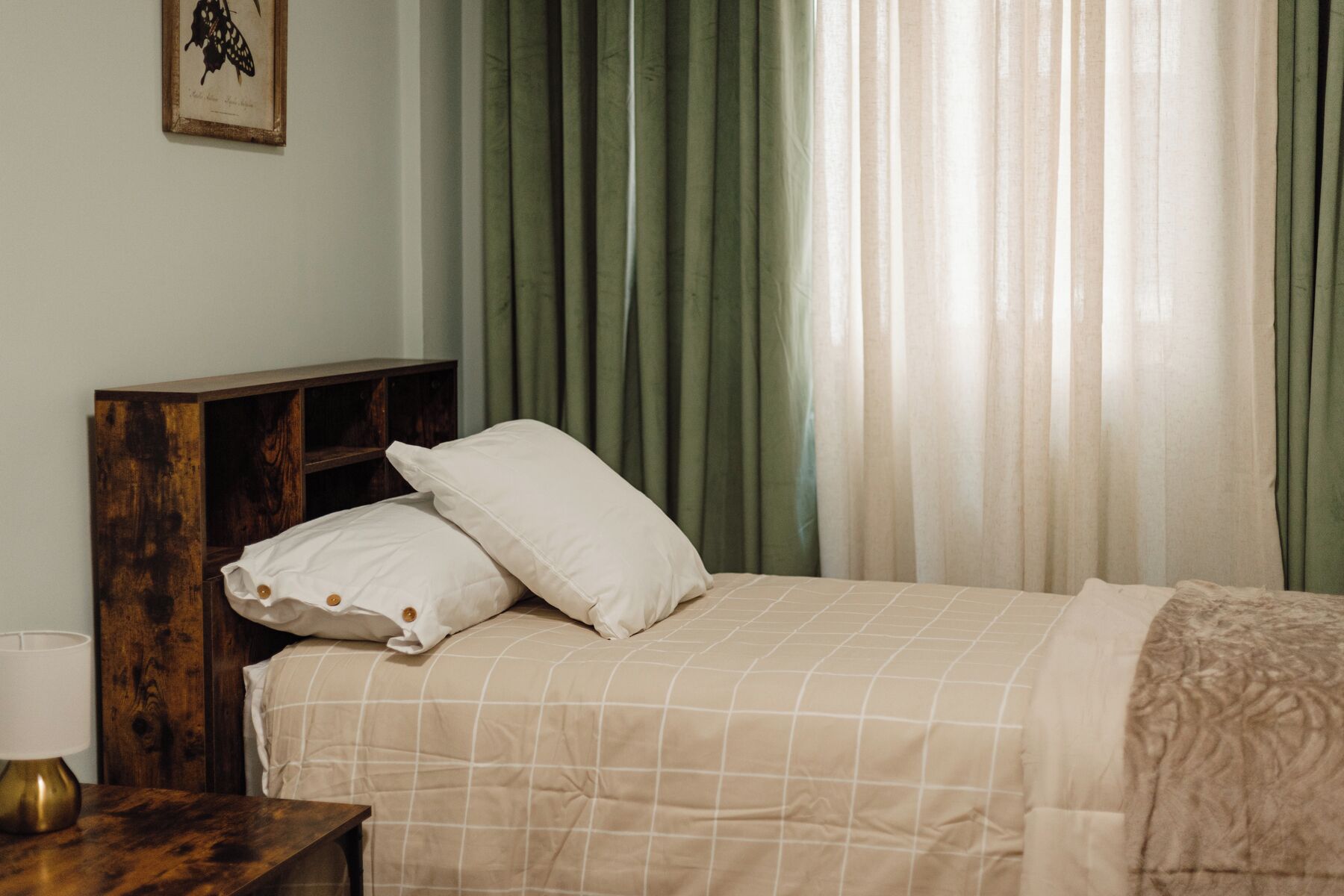 Una cama bien hecha con una colcha blanca y color beige a cuadros, dos almohadas blancas y una mesita de noche de madera con una lámpara.