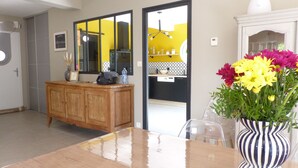 A dining area with a wooden table, chairs, a vase with flowers, and a kitchen visible through an open door.