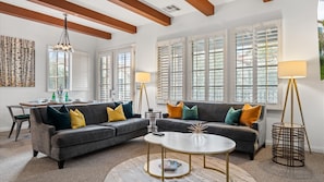 A living room with a sectional sofa, a round coffee table, a dining table with chairs, and large windows with shutters.