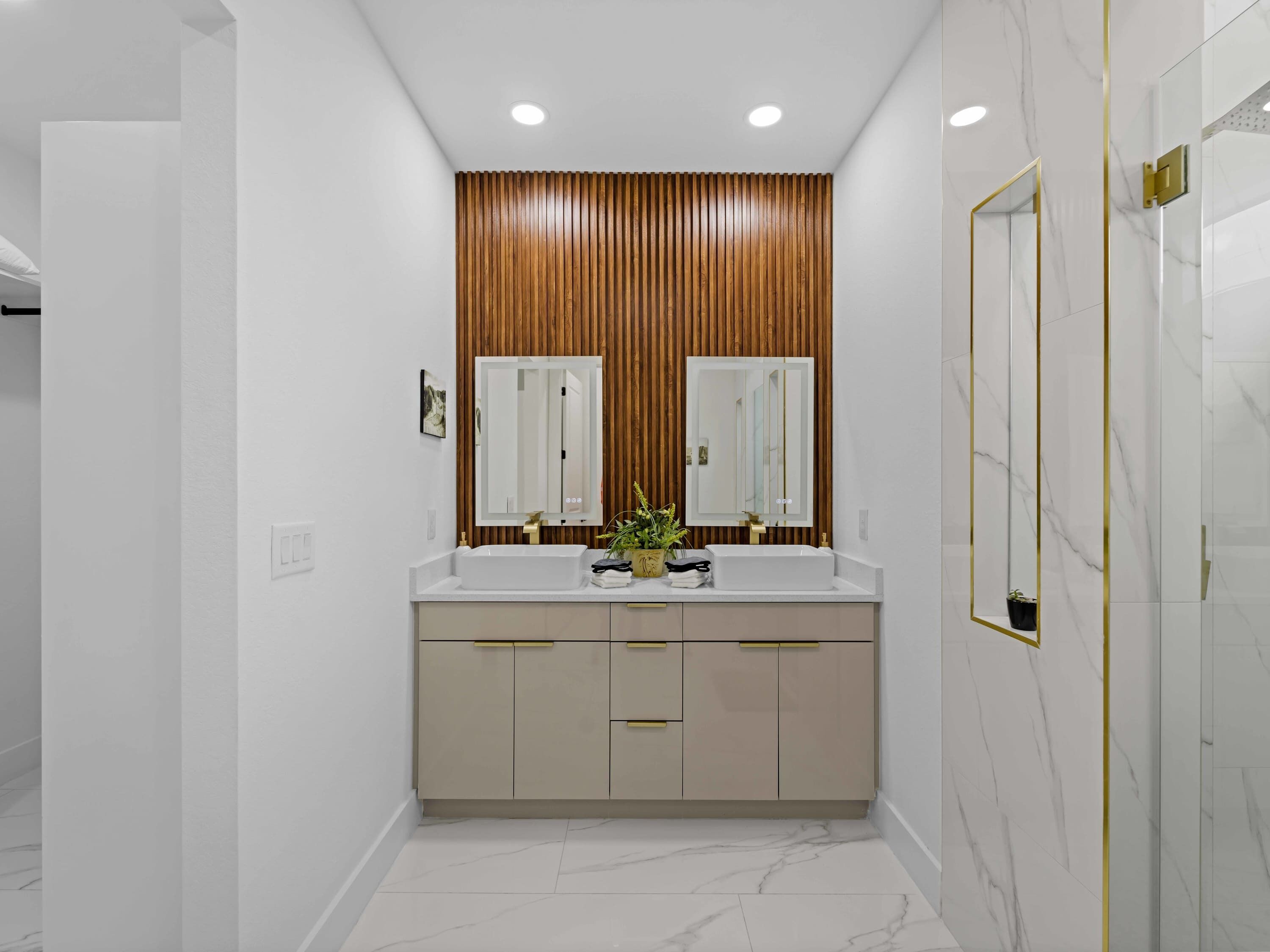 Un baño moderno con dos lavamanos, pared de paneles de madera, suelo de mármol y espejos con marco dorado.