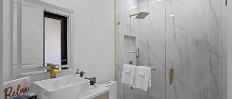 Un bagno moderno con lavandino bianco, accessori dorati, cabina doccia in vetro e pareti in marmo.
