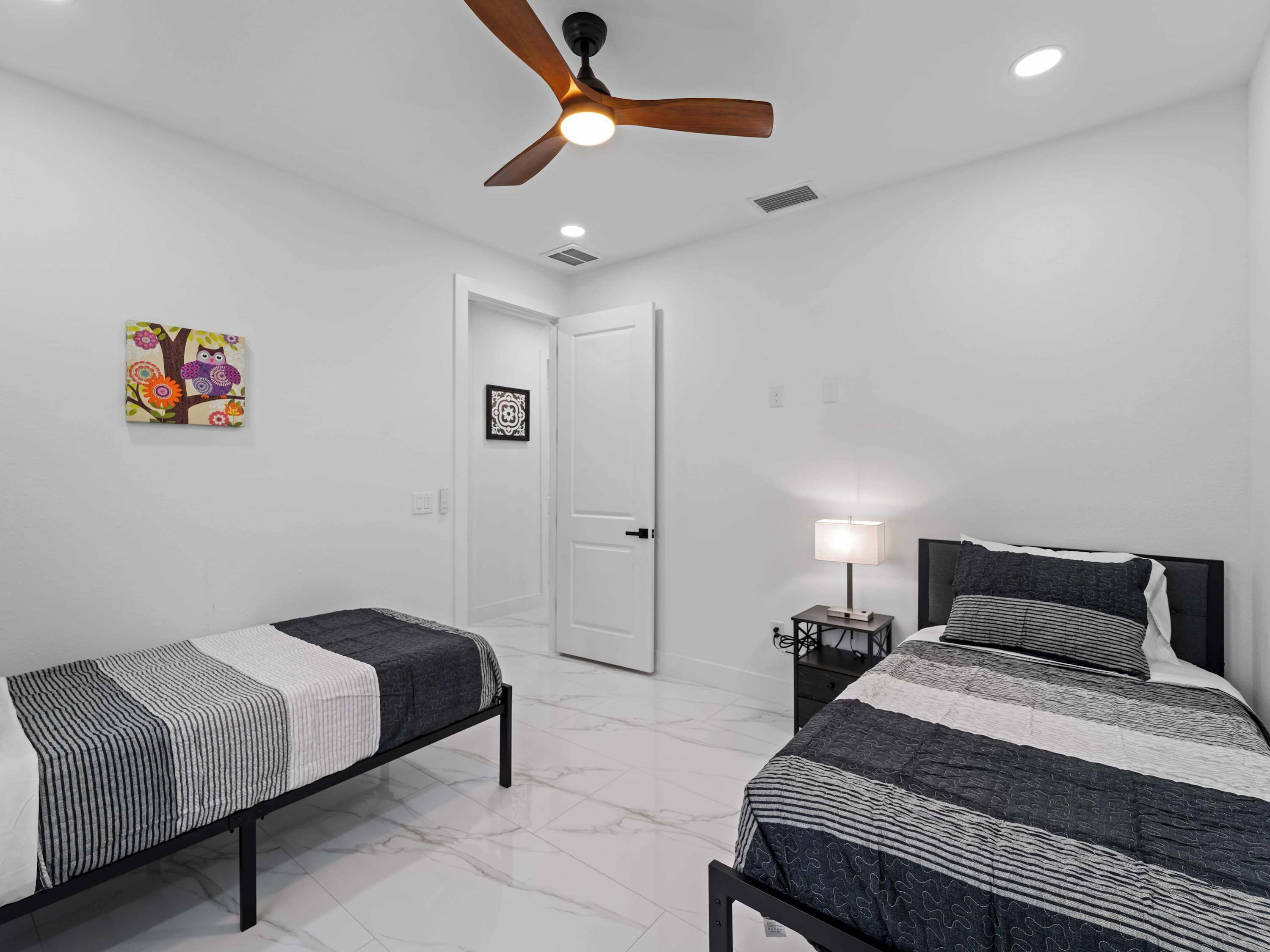 A bedroom with two beds, a ceiling fan, a nightstand with a lamp, and a door.