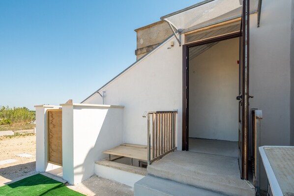 A modern building with a staircase leading to a door, a small outdoor area with a green mat, and a clear sky.