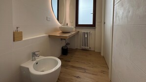 Un baño moderno con un lavabo blanco, un bidé, un mueble de tocador de madera, un espejo y una ventana.