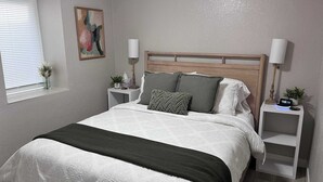 A neatly made bed with a white and green bedspread, two bedside tables with lamps, a small plant, and a painting on the wall. - Duluth Delight #1 Sum (Sioux Falls)