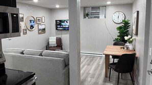 A modern living room with a sofa, a dining table with chairs, a TV mounted on the wall, and a clock. - Duluth Delight #1 Sum (Sioux Falls)