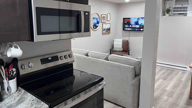 A modern kitchen with a built-in microwave, oven, and stovetop. There is a grey sofa in the background, a flat-screen TV mounted on the wall, and a small potted plant. - Duluth Delight #1 Sum (Sioux Falls)