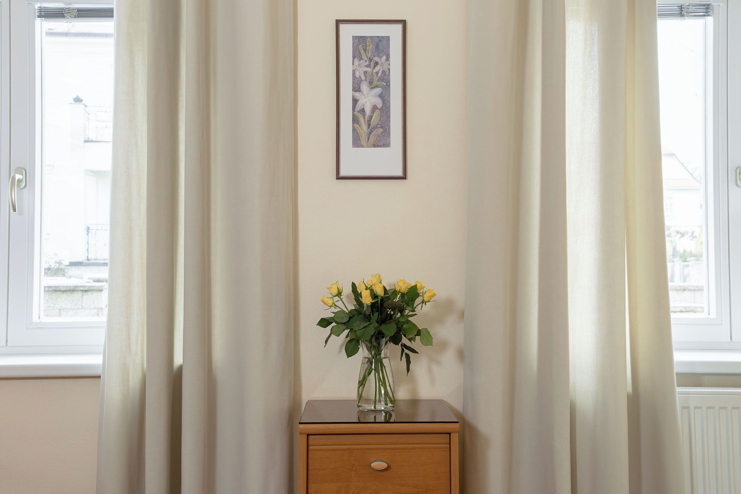 A room with a wooden nightstand, a vase with yellow flowers, and a framed picture on the wall.