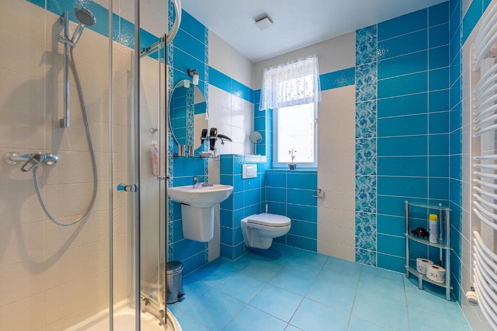 A bathroom with blue tiles, a glass shower enclosure, a sink, a toilet, and a window with a white curtain.