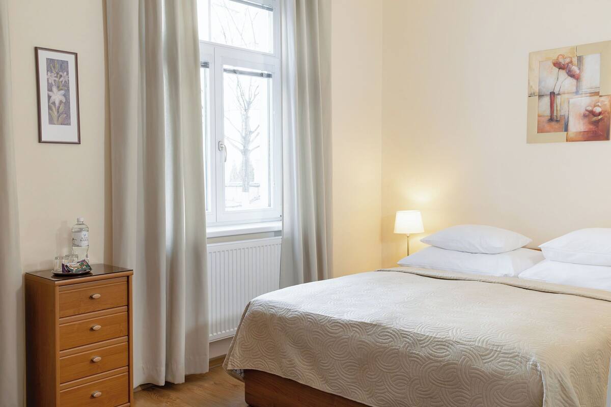 A bedroom with a bed, a nightstand, a window with curtains, and a wall-mounted lamp.