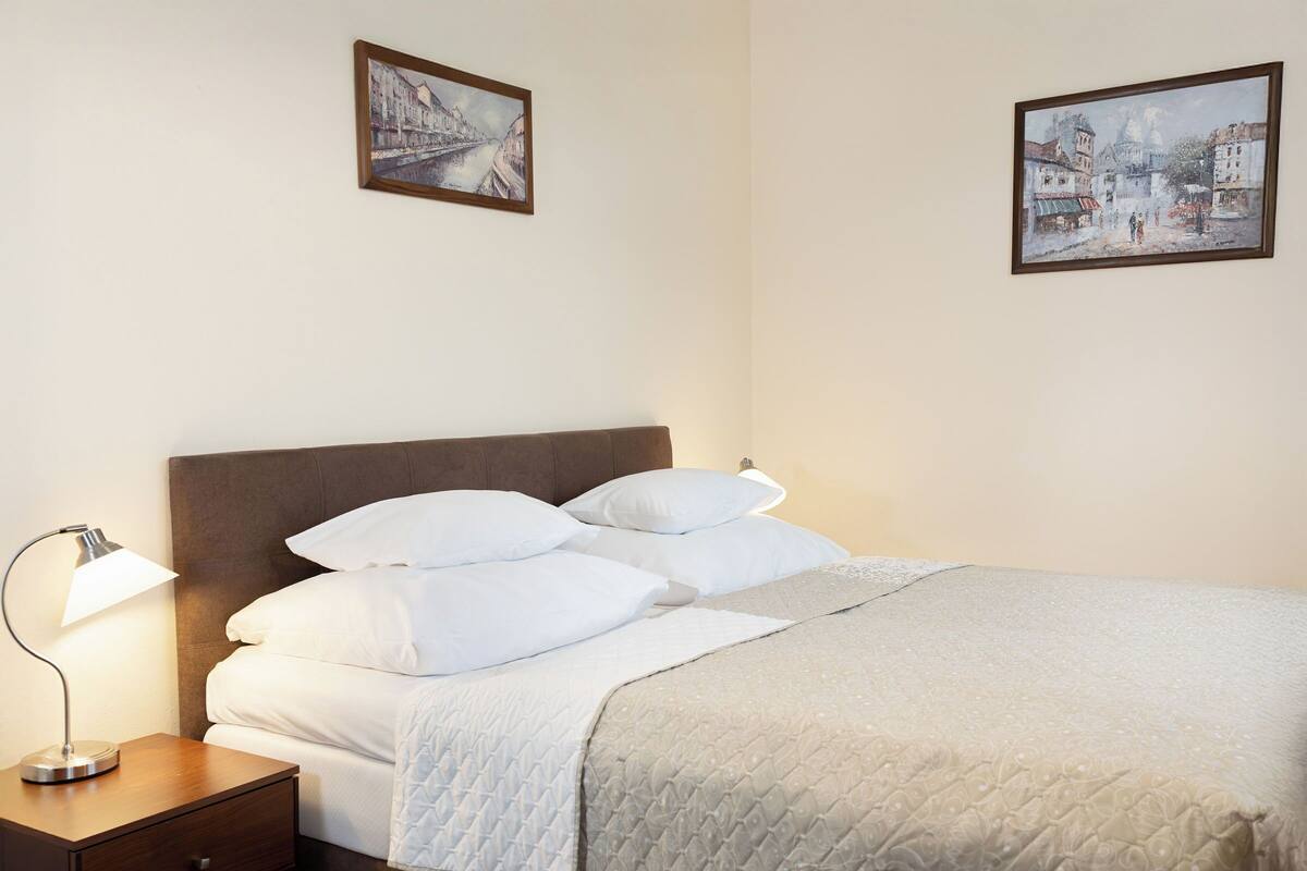 A neatly made bed with white linens and a quilted blanket, a bedside lamp, and two framed paintings on the wall.