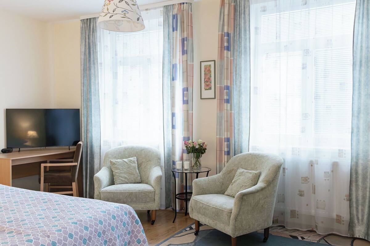 A hotel room with a bed, two armchairs, a desk with a TV, and a window with curtains.