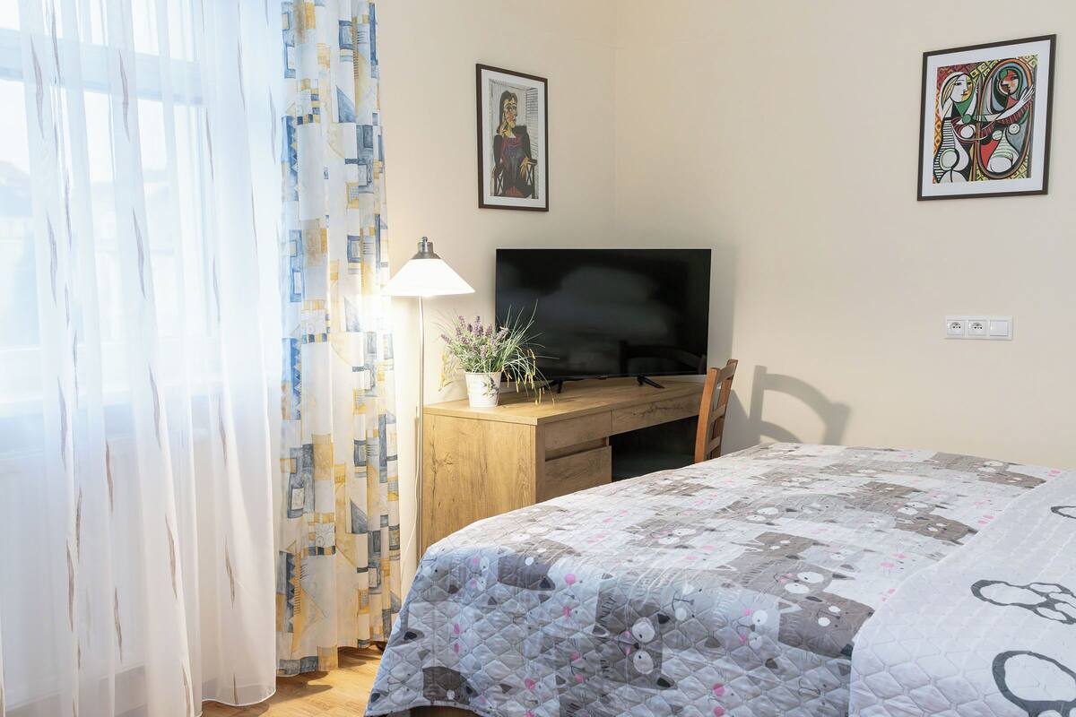 A bedroom with a bed, a desk with a TV, and two framed artworks on the wall.