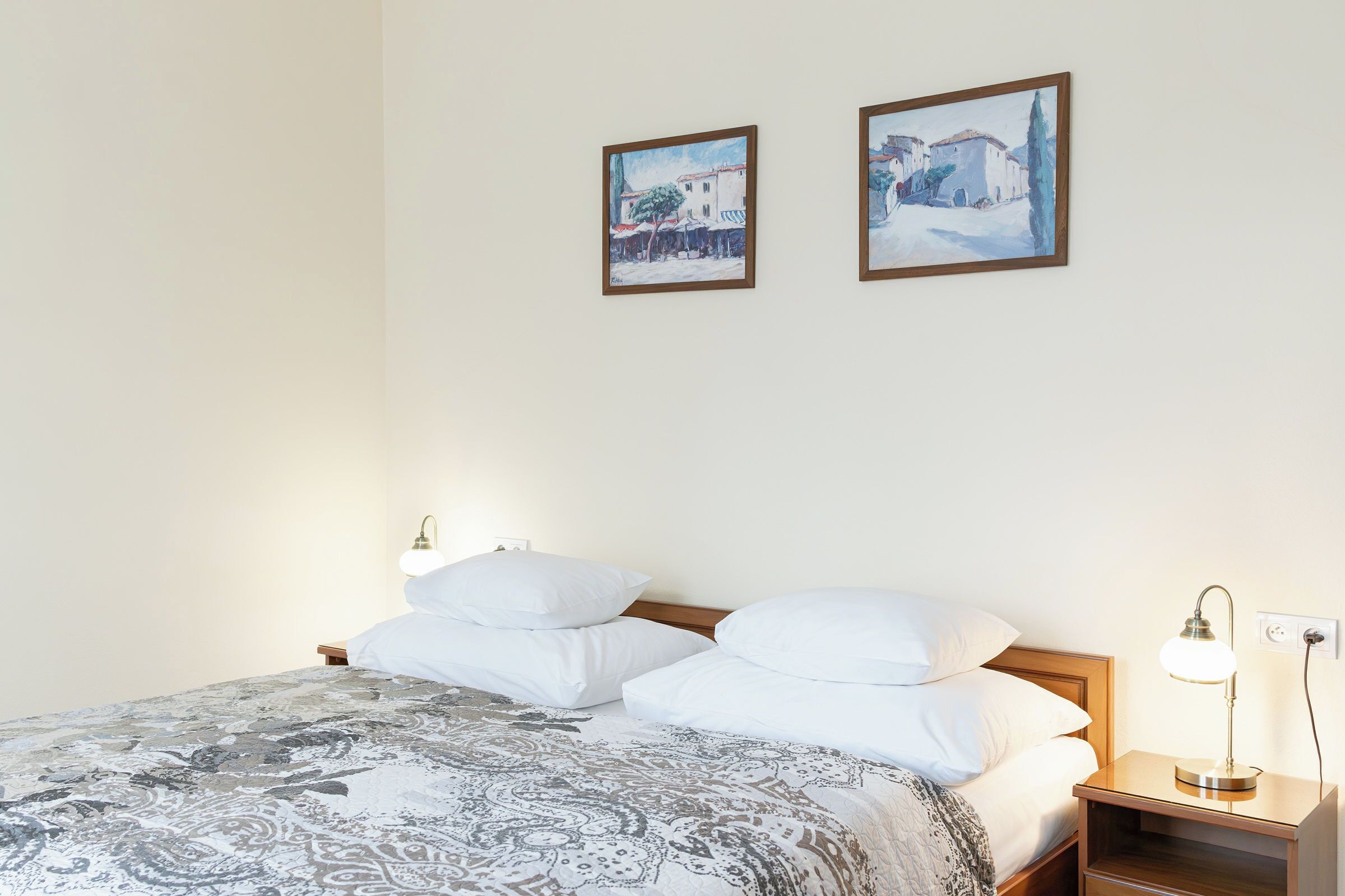 Una cama bien hecha con una colcha estampada, dos almohadas blancas, una mesita de noche con una lámpara y dos cuadros en la pared.