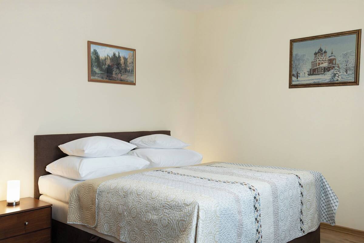 A neatly made bed with a patterned bedspread, a wooden nightstand with a lamp, and two framed pictures on the wall.