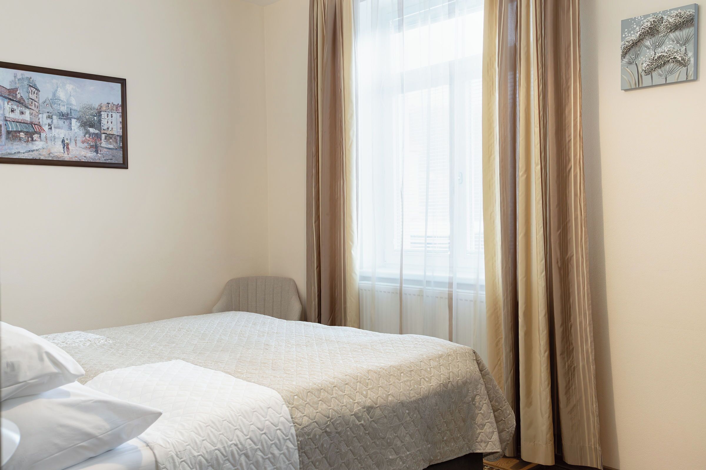 Una habitación de hotel con cama, una silla, ventana con cortinas y un cuadro en la pared.