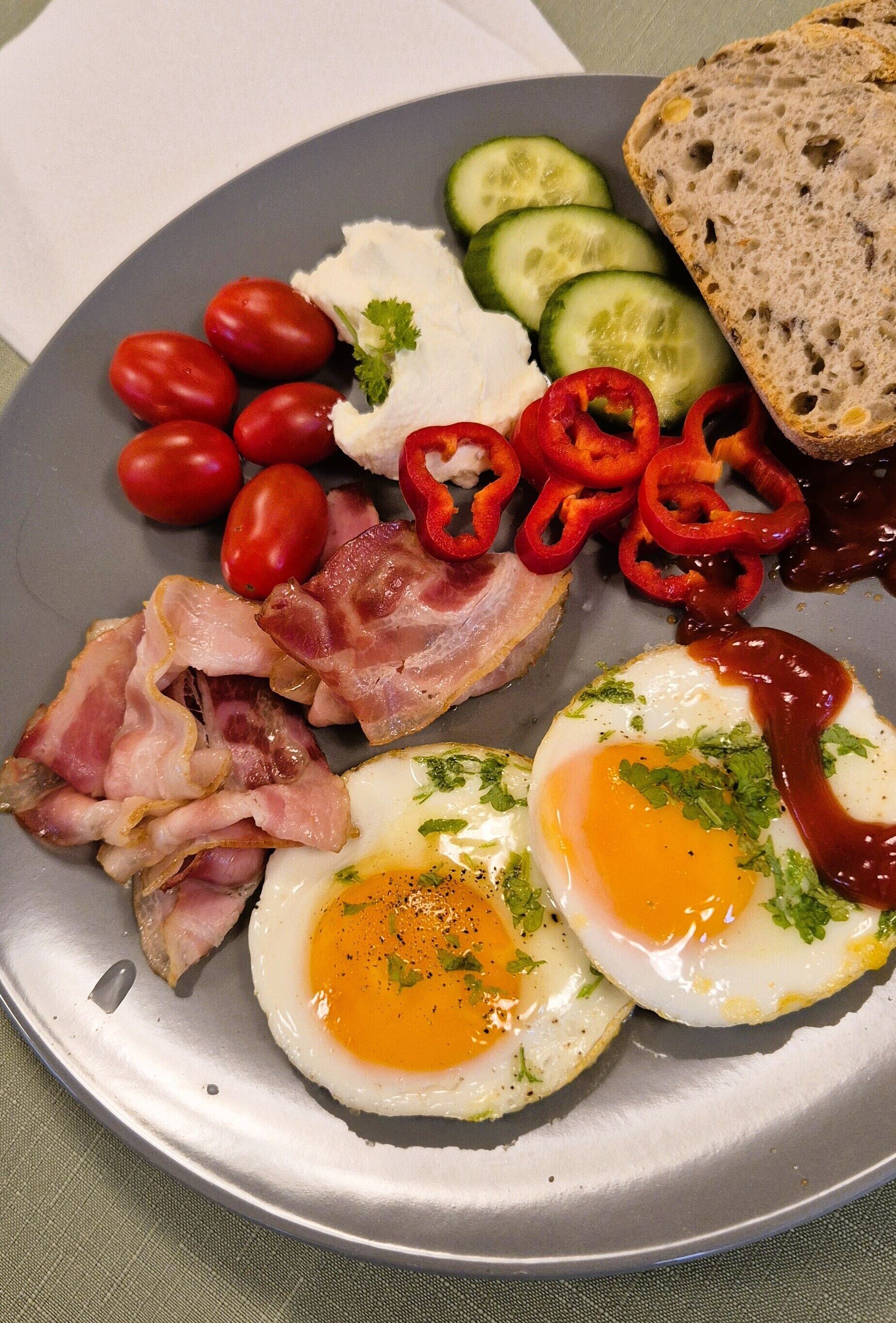 Une assiette contenant des œufs, du bacon, des tomates, des concombres, des poivrons rouges et du pain.