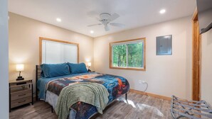 Un dormitorio con cama, mesitas de noche, ventilador de techo y ventana con vista a árboles.