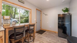 Cocina compacta con una mesa de madera, dos taburetes, un refrigerador y una ventana con vistas a un vehículo y follaje.