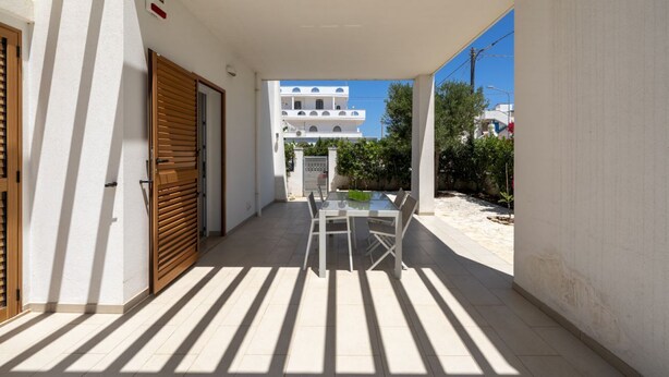 Zona exterior coberta amb taula i cadires, una porta de fusta i una vista d'un edifici blanc al fons.