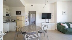 Cuina moderna amb armaris blancs, microones i nevera. Hi ha una zona de menjador amb una taula rodona i cadires. Un televisor de pantalla plana està fixat a la paret.