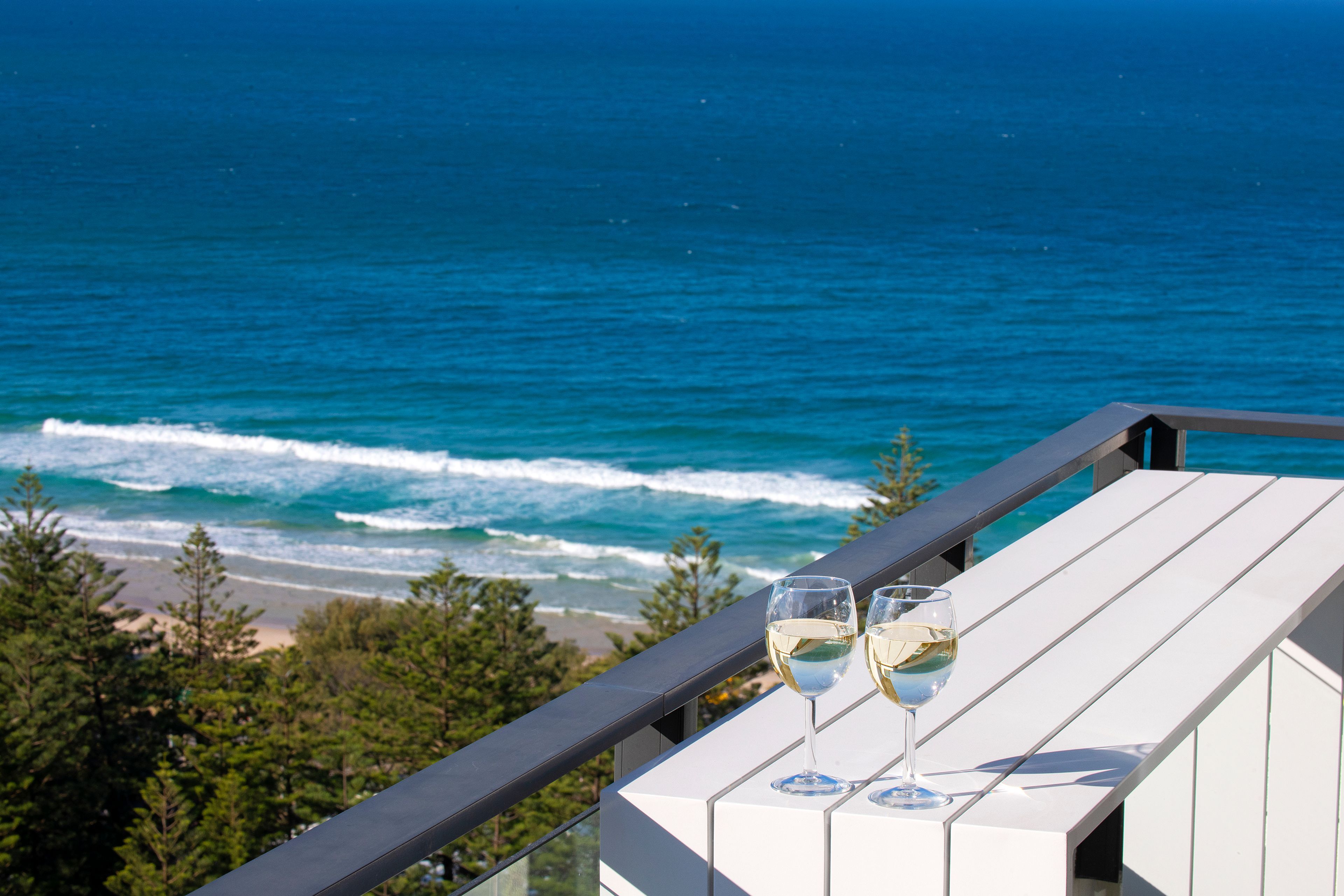 A balcony with two glasses of white wine, overlooking a beach and ocean.